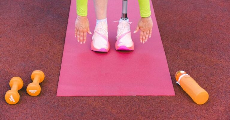 Practices - Unrecognizable female athlete with leg prosthesis bending forward and stretching while trying ti touch ground during training on sports ground with dumbbells